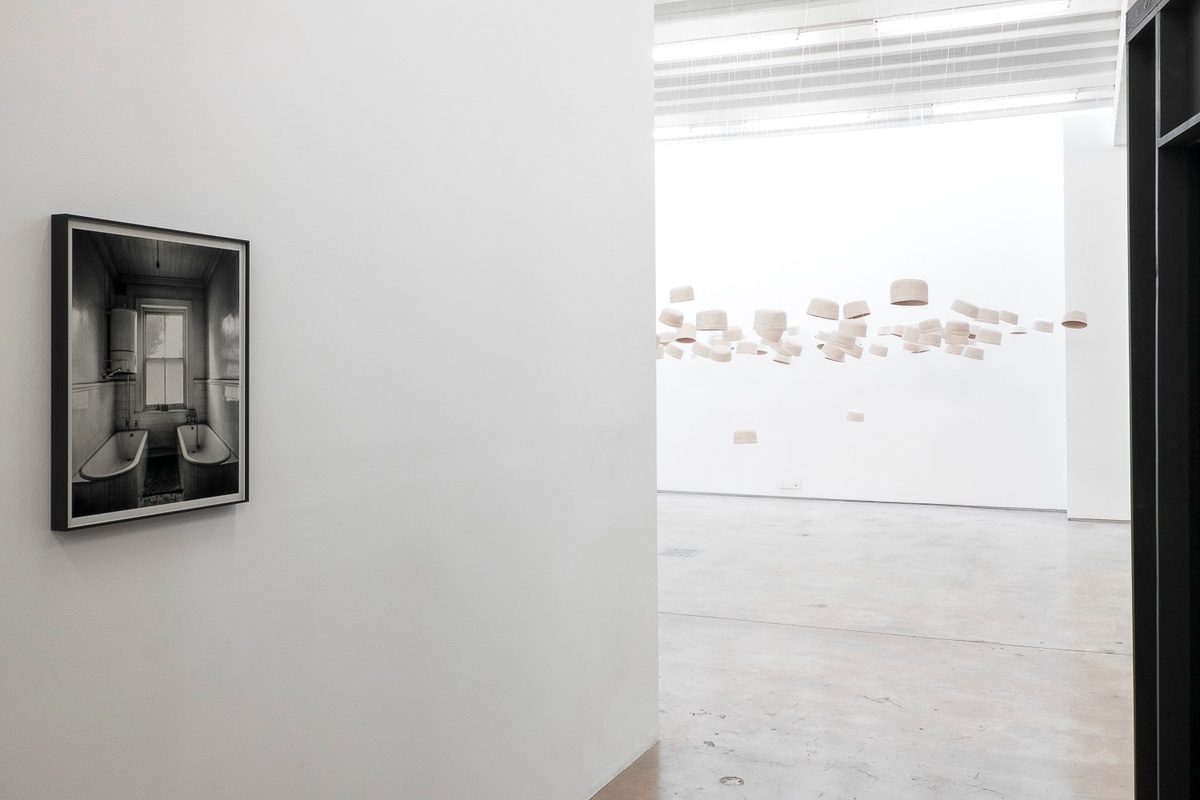 Installation photograph from the You & I exhibition in A4’s Gallery. On the left, David Goldblatt’s monochrome photograph ‘For the General Manager after he had been underground: 'dirty' bath and "clean" bath attached to the General Manager's office, New Kleinfontein Gold Mine, Benoni. May 1967’ is mounted on the gallery wall. On the right, casts of kufiya’s from Haroon Gunn-Salie and James Matthews’ installation ‘Amongst Men’ are suspended from the ceiling.
