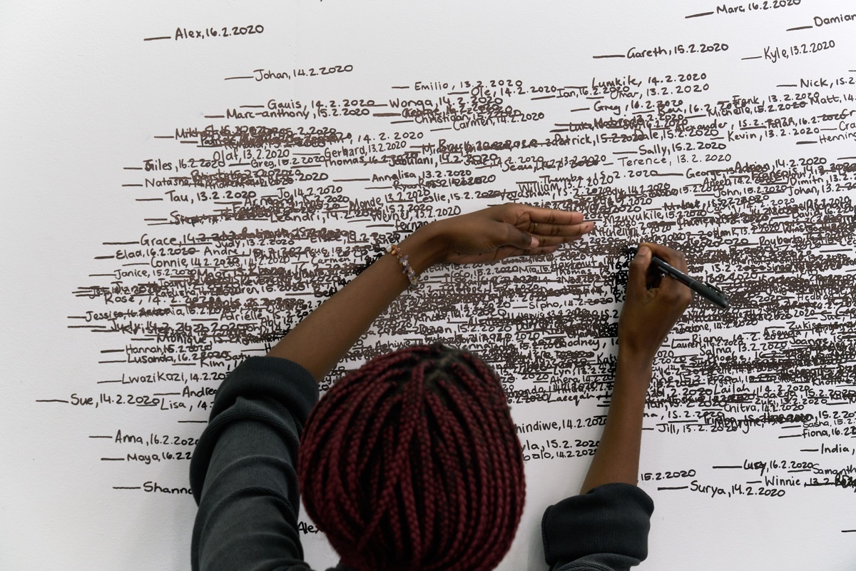 Event photograph of Roman Ondák’s performance piece ‘Measuring the Universe’ at A4’s booth at the 2020 Cape Town Art Fair. At the back, a white wall is covered with overlapping marks that indicate participant’s height, names and the date of their participation in black felt pen. At the front, an A4 facilitator ads information to the wall.
