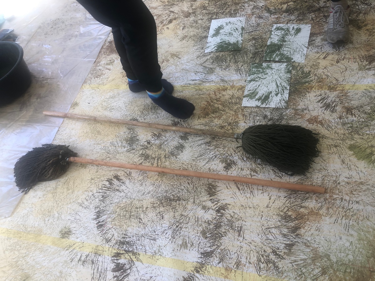 Process photograph from ‘Open Production’, Igshaan Adams’ hybrid studio/exhibition in A4’s Gallery. A topdown view shows the floor lined with paper covered in marks made using a mop, with two mops lying on top of it.
