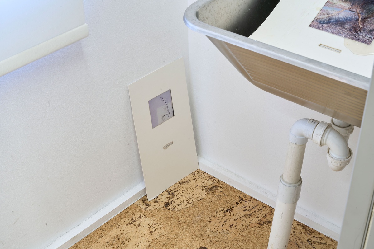 Installation photograph of Michael Tymbios’ studio during his residency in A4 Arts Foundation. On the left, a piece of backing board with a photographic print leans against the base of a wall. On the right, a piece of backing board with a photographic print sits in a metal sink.
