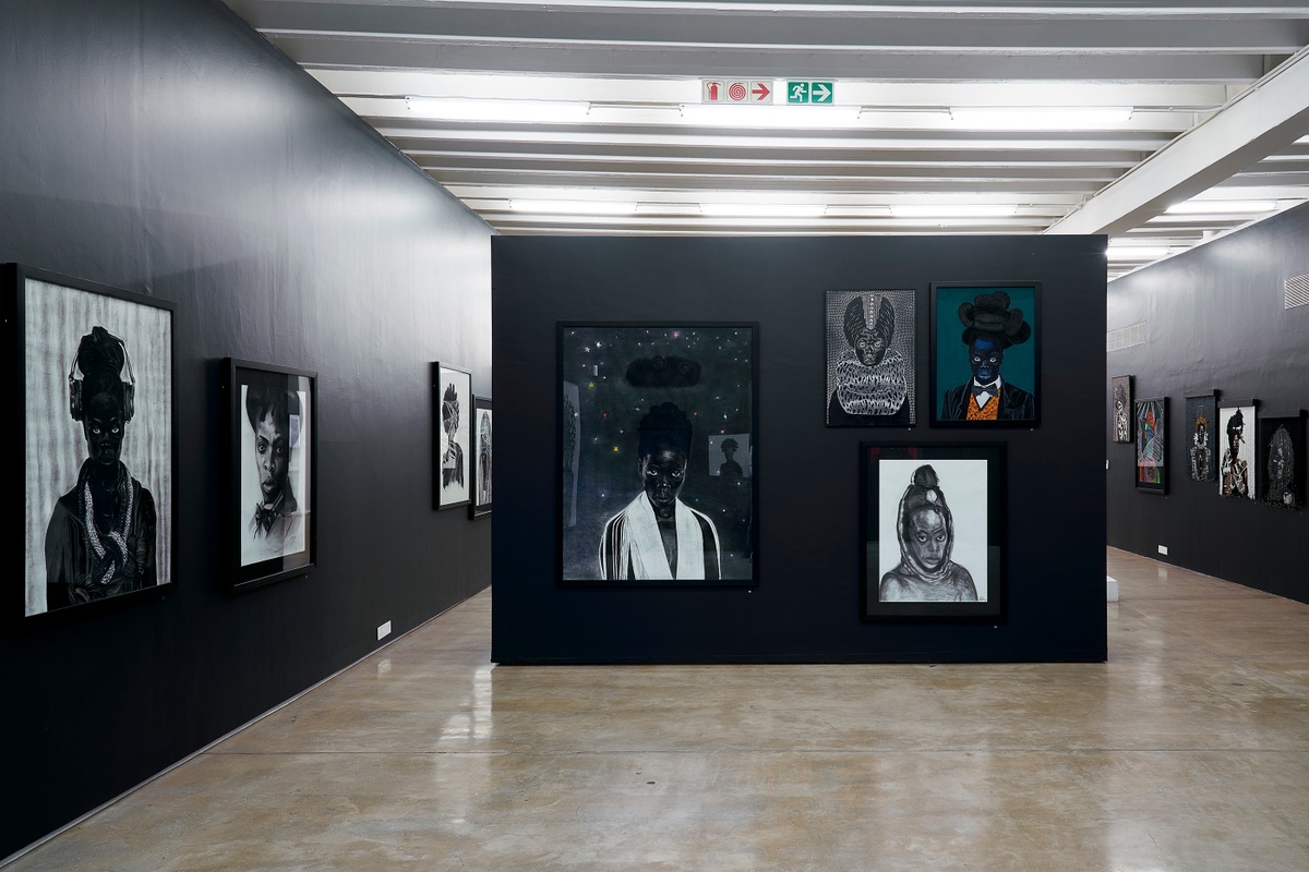 Installation photograph from the “Ikhono LaseNatali” exhibition in A4’s Gallery that shows a black moveable gallery wall. On the left, Lindani Nyandeni’s woodcut “Somnyama Ngonyama, ‘Zinathi I’” is mounted to the wall. On the right, Sthenjwa Luthuli’s woodcut “Somnyama Ngonyama ‘Somnyama III’,” Andile Maphumulo’s acrylic painting “Somnyama Ngonyama” and Londi Mtshali’s charcoal drawing “Somnyama Ngonyama, ‘Ntombi I’” are mounted to the wall.
