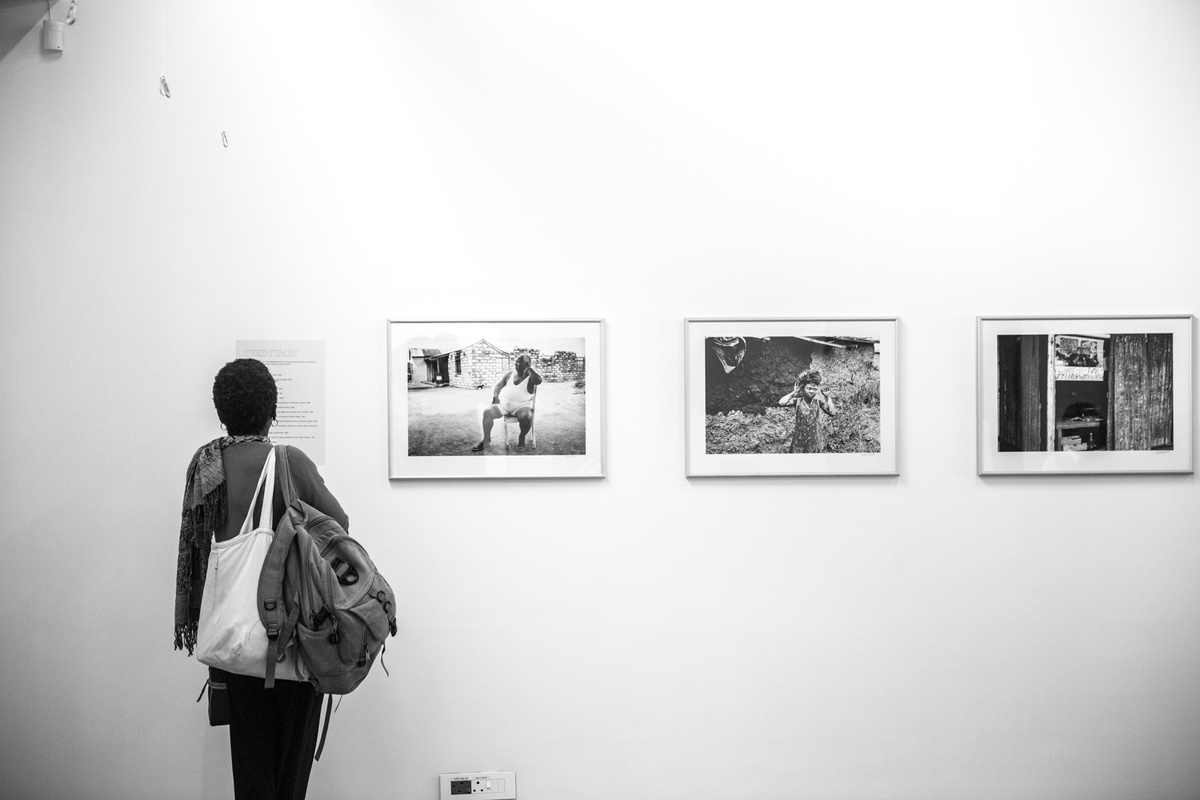 Monochrome event photograph from the launch of Omar Badsha’s book ‘Seedtimes’ on A4’s top floor. At the back, a printed list of works and Omar Badsha’s monochrome photographs ‘Induna Nkonyane, Amouti, Kwazulu-Natal’, ‘Young woman repairing home, Inanda, Kwazulu-Natal’ and ‘Watch repair shop, Inanda, Kwazulu-Natal’ are mounted on the wall. At the front, an attendee reads the list of works.
