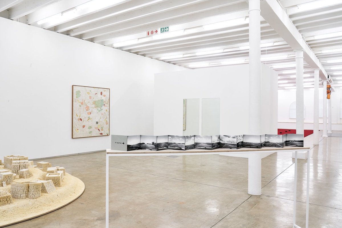 Installation photograph from The Future Is Behind Us exhibition in A4’s Gallery. At the front, Jo Ractliffe’s open-spine accordion book ‘N1: every 100 kilometers’ sits on a thin rectangular table. On the left, Kader Attia’s couscous sculpture from ‘Untitled (Ghardaïa)’ resembles a town. At the back, Asemahle Ntlonti’s framed mixed media work ‘Emaphandleni’ hangs on the wall on the left, and Félix González-Torres’ mirror installation '“Untitled” (Orpheus, Twice)' is mounted on a moveable gallery wall on the right.
