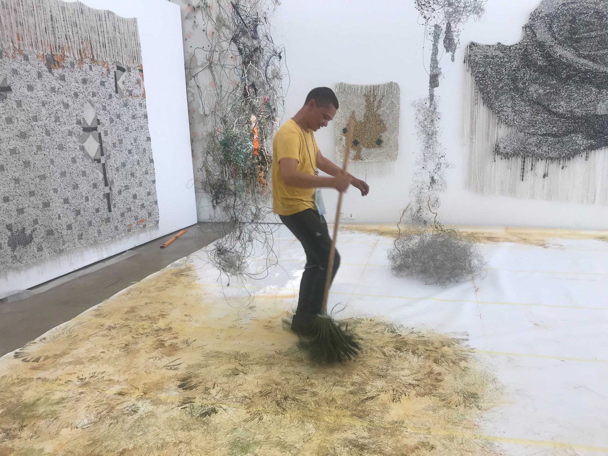 Process photograph from ‘Open Production’, Igshaan Adams’ hybrid studio/exhibition in A4’s Gallery. At the back, two of Adams’ mixed media wire sculptures are suspended from the ceiling. At the front, Adams makes marks on the paper lining the gallery floor with a mop.
