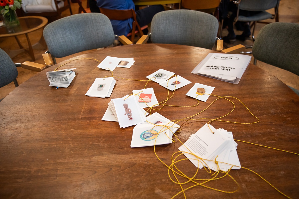 Event photograph from the preview of the 'A Little After This' exhibition in A4 Arts Foundation shows the printed index cards for the collected objects from Penny Siopis' 'Will' work on a wooden table. The cards feature eyelets and are threaded together to form an expandable book.
