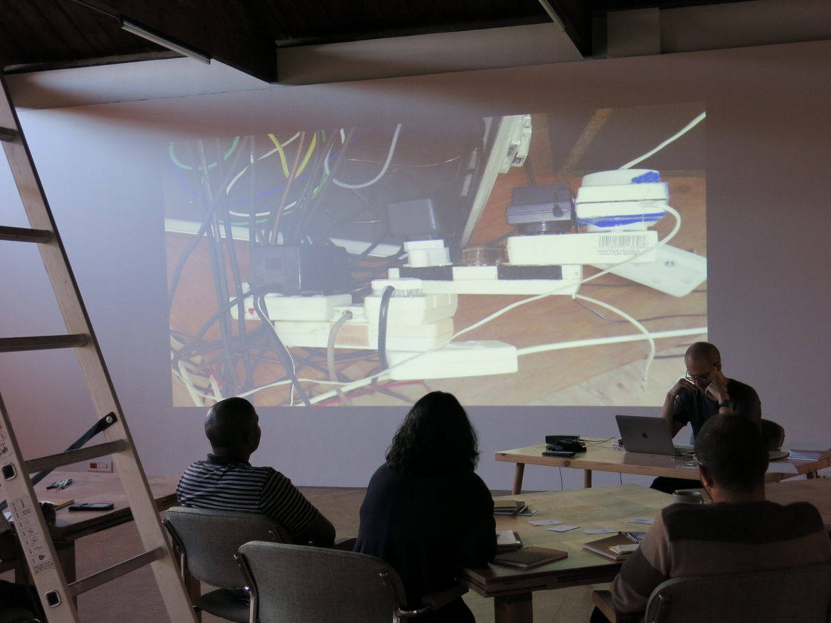 Event photograph from the 2018 rendition of the City Research Studio exchange with the African Centre for Cities. At the front, participants and contributors are seated at wooden tables arranged along the room. At the back, a wall projected photograph of stacked power adaptors.
