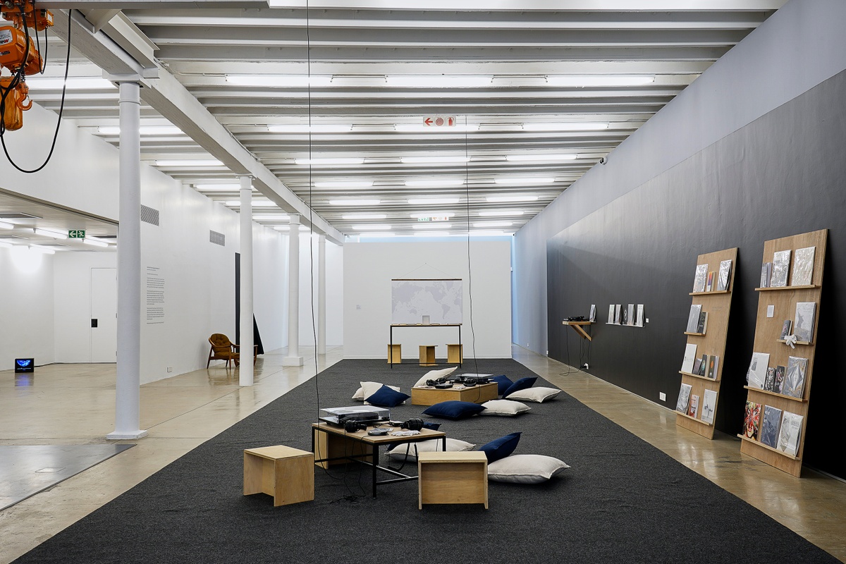 Installation photograph from the ‘Sounding the Void, Imaging the Orchestra V.1’ exhibition in A4’s Gallery. At the front, two small low tables with vinyl record players and headphones. At the back, Andrew Pekler’s print media and sound installation ‘Phantom Islands - A Sonic Atlas’. On the right, a selection of books and vinyls from Bhavisha Panchia’s ‘Nothing to Commit Records’ sits on two wooden shelves and a selection of books and cassettes from the Sahel Sounds label sit on wall-mounted shelves.
