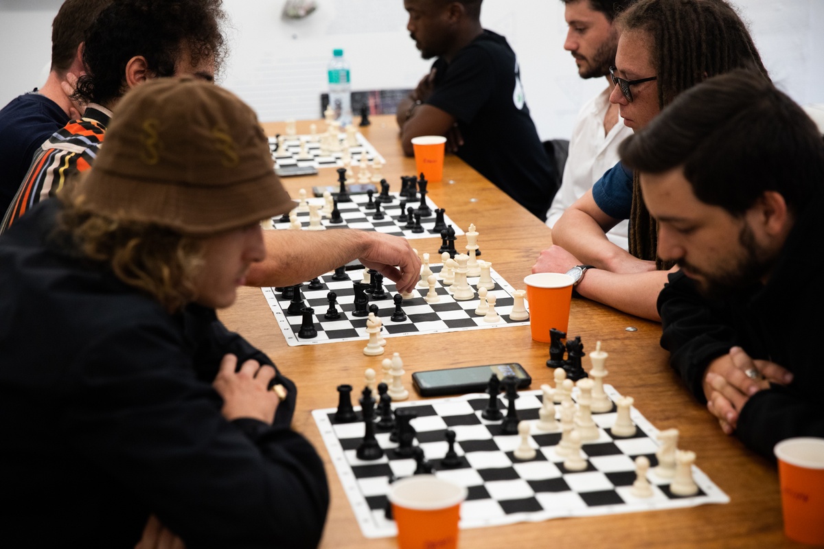 Event photograph from the launch of Brett Seiler’s chess set in A4’s Proto museum shop. A long wooden table features four travel chess sets, each with a pair of competitors.
