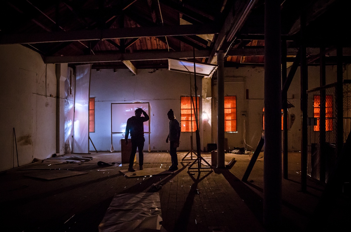 Process photograph from George Mahashe’s residency on A4’s top floor. Three freestanding blank surfaces are arranged around the space so as to catch refracted light from A4’s exterior through Mahashe’s pinhole cameras. At the front, two individuals are standing silhouetted in the darkened space.
