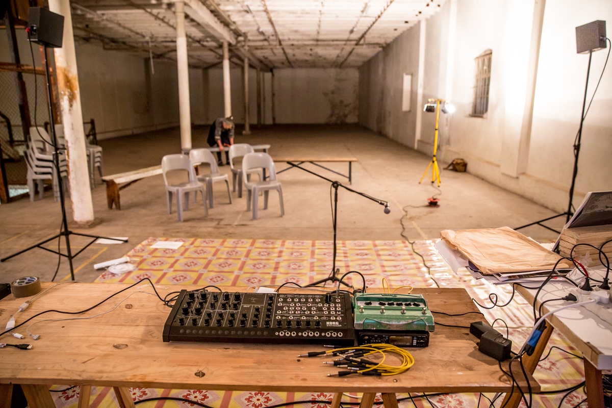 Process photograph from the ‘Future Nostalgia’ event with musician João Orecchia on A4’s first floor. At the front, a wooden table with an audio mixer. At the back, a performance area covered with laminated flooring, and seating for audience members being arranged.
