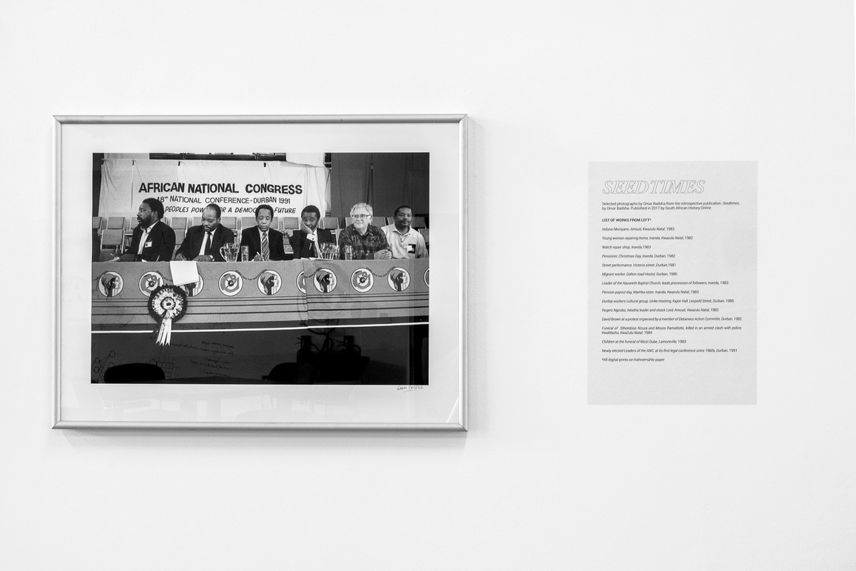 Installation photograph from the launch of Omar Badsha’s book ‘Seedtimes’ on A4’s top floor. On the left, Omar Badsha’s monochrome photograph ‘Newly elected leaders of the ANC at its first legal conference since 1960's, Kwazulu-Natal’ is mounted on the gallery wall. On the right, a list of works on a page is mounted on the gallery wall.
