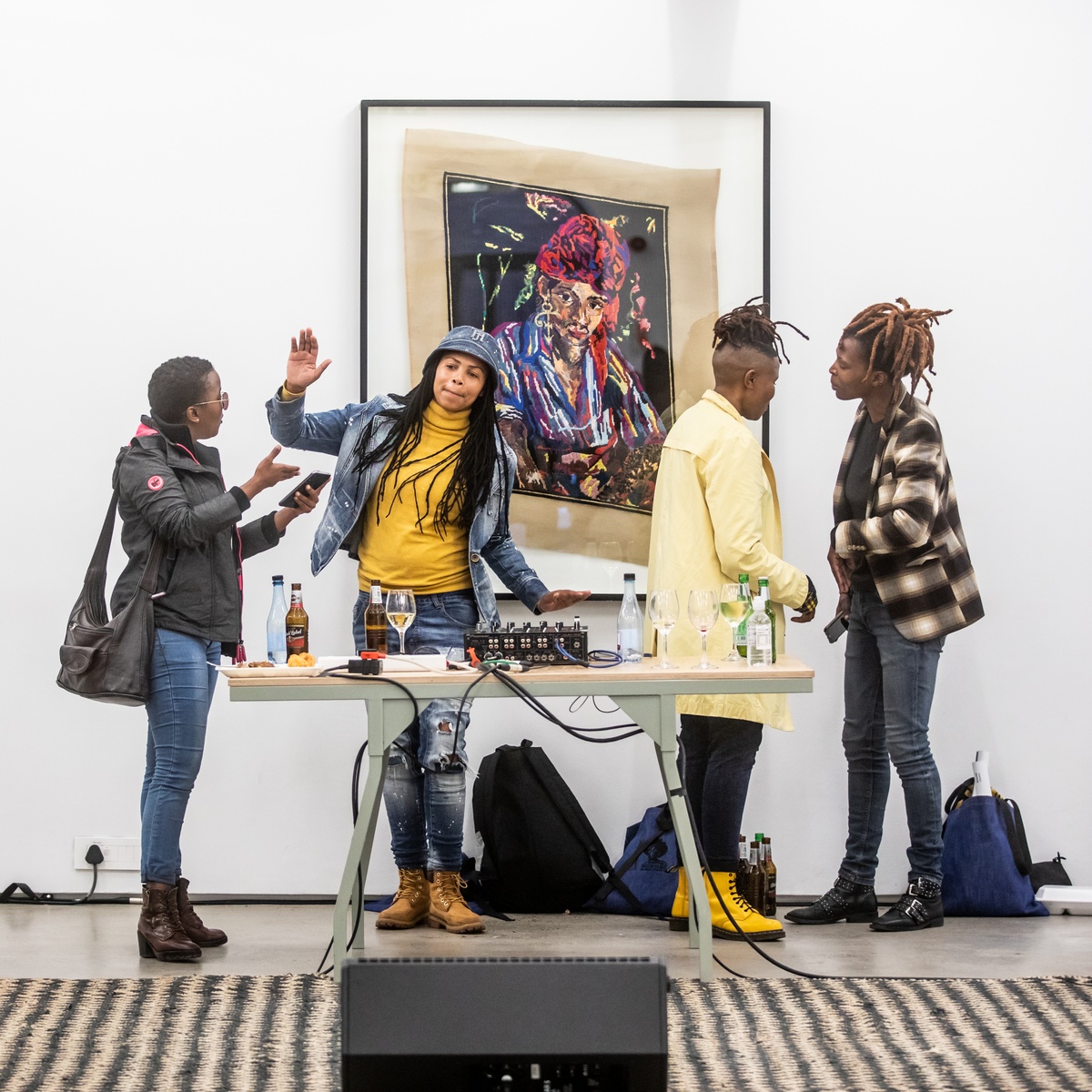 Event photograph from the opening of the “Ikhono LaseNatali” exhibition in A4’s Gallery. At the back, Athi-Patra Ruga’s crocheted work “Arab Boy (after Irma Stern)” is mounted on the gallery wall. At the front, four individuals stand behind a desk with audio equipment.

