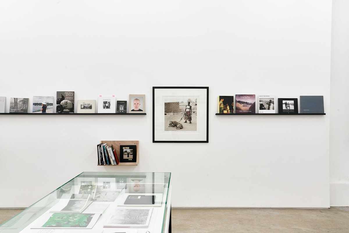 Installation photograph from the Photo Book! Photo-Book! Photobook! exhibition in A4’s Gallery. At the front, a freestanding glass display case with printed matter. At the back, shelving units with printed matter from the years 1994 to 2022 line a white wall, along with Pieter Hugo’s photograph ‘Mallam Mantari Lamal with Mainasara, Abuja, Nigeria, 2005’.
