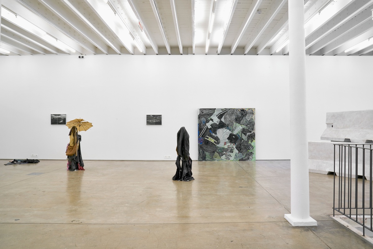 Installation photograph from the ‘without a clear discernible image’ exhibition in A4’s Gallery. On the left, Kevin Beasley’s untitled resin, garment and umbrella sculpture stands on the gallery floor. In the middle, Beasley’s resin, garment and walking stick sculpture ‘Walker’ stands on the gallery floor. At the back, Beasley’s monochrome photographs ‘A view of the 12 Apostles’ and ‘Robot’ are mounted on the wall, and his resin, garment and metal sculpture ‘California South Africa’ sits at the base of the wall. 
