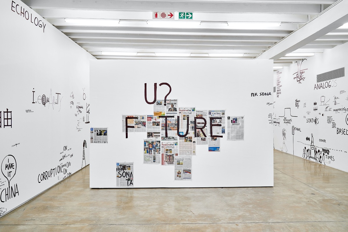 Installation photograph from Dan Perjovschi’s ‘The Black and White Cape Town Report’ exhibition in A4’s Gallery. In the middle, a moveable gallery wall features a collage of newspaper articles overlain with the word ‘future’ in black felt pen marker, where the first ‘u’ is elevated above the other letters and accompanied by a question mark.
