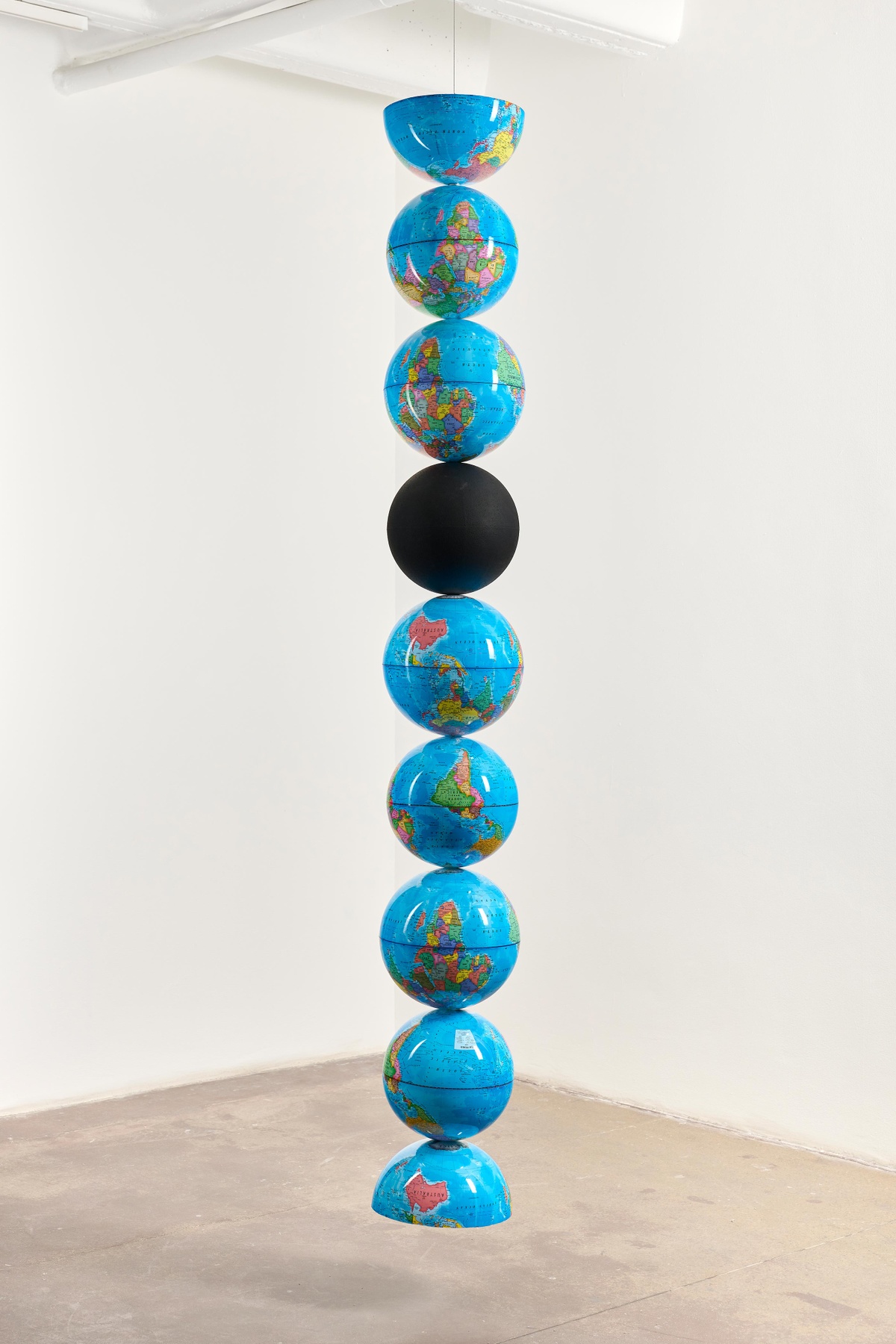 Installation photograph that depicts Nolan Oswald Dennis’ ‘model for an endless column’ a column of PET globe models, suspended from a ceiling. One is solid black, the first and last are halved.

