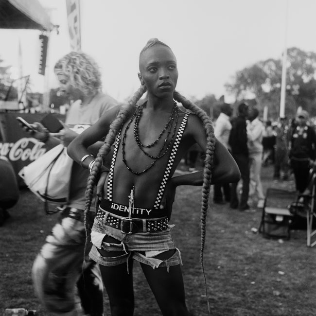 Monochrome photograph ‘Identity’ from Sabelo Mlangeni’s residency on A4’s top floor. At the front, a person standing in a grassy area. At the back, a crowd is gathered next to a stage.
