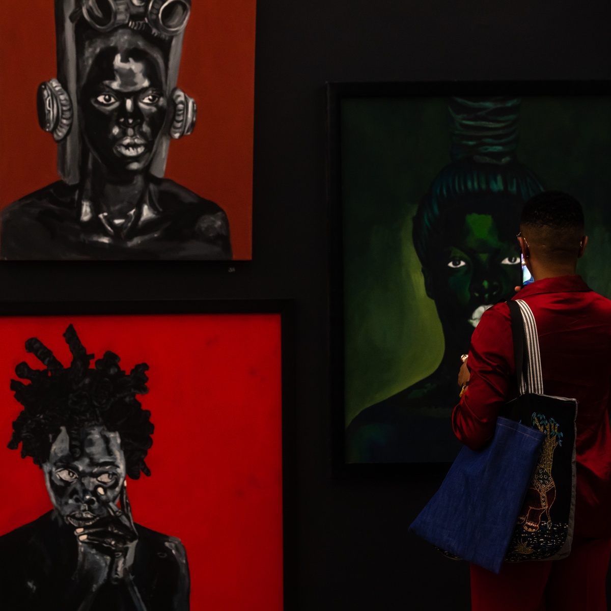 Event photograph from the opening of the “Ikhono LaseNatali” exhibition in A4’s Gallery. At the back, Ncumisa Mcitwa’s acrylic painting “Somnyama Ngonyama, ‘Namhla at Cassibaus’,” Sphephelo Mnguni’s acrylic painting “Somnyama Ngonyama ‘Khwezi’” and Nomusa Musah Mtshali’s acrylic painting “Somnyama Ngonyama” are mounted on a black gallery wall. At the front, an attendee stands in the gallery.
