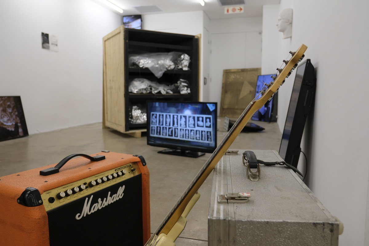 Process photograph from NRNA’s launch of their EP ‘Kalahari Elegy’ on A4’s top floor. At the front, an electric guitar and amplifier sit on the gallery floor. At the back, process work from Rodan Kane Hart’s ‘Western Death Mask(s)’ project, which includes a screen on the floor displaying images of busts and a wooden crate filled with stainless steel sculptures.
