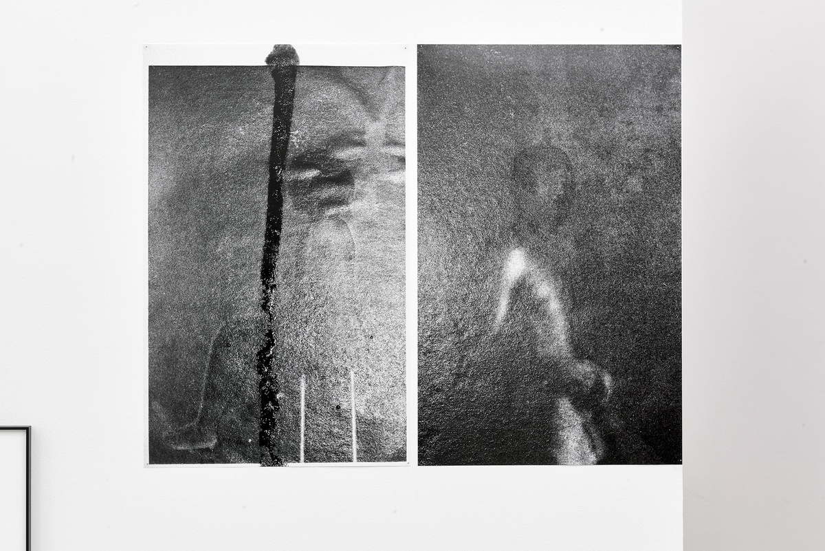 Two grainy black and white photographs, hanging side by side. The left-hand photo shows abstract lines and shapes. The right-hand side photo is of a figure from the waist up, partly obscured.
