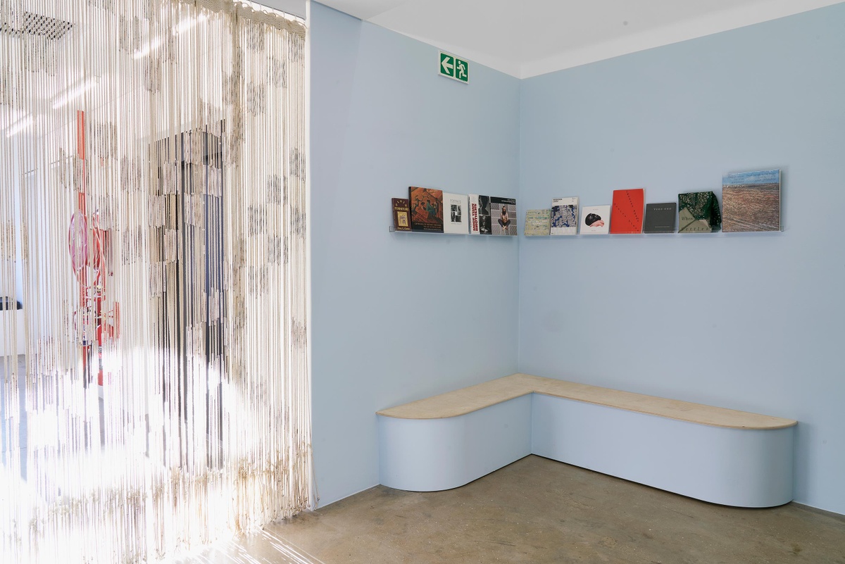 Installation photograph from the Customs exhibition in A4’s Gallery. On the left, Igshaan Adams’ tapestry ‘Al asma ul Husna’ is suspended from the ceiling. On the right, a wall-mounted shelf holds a selection of printed materials above a seating area for reading.
