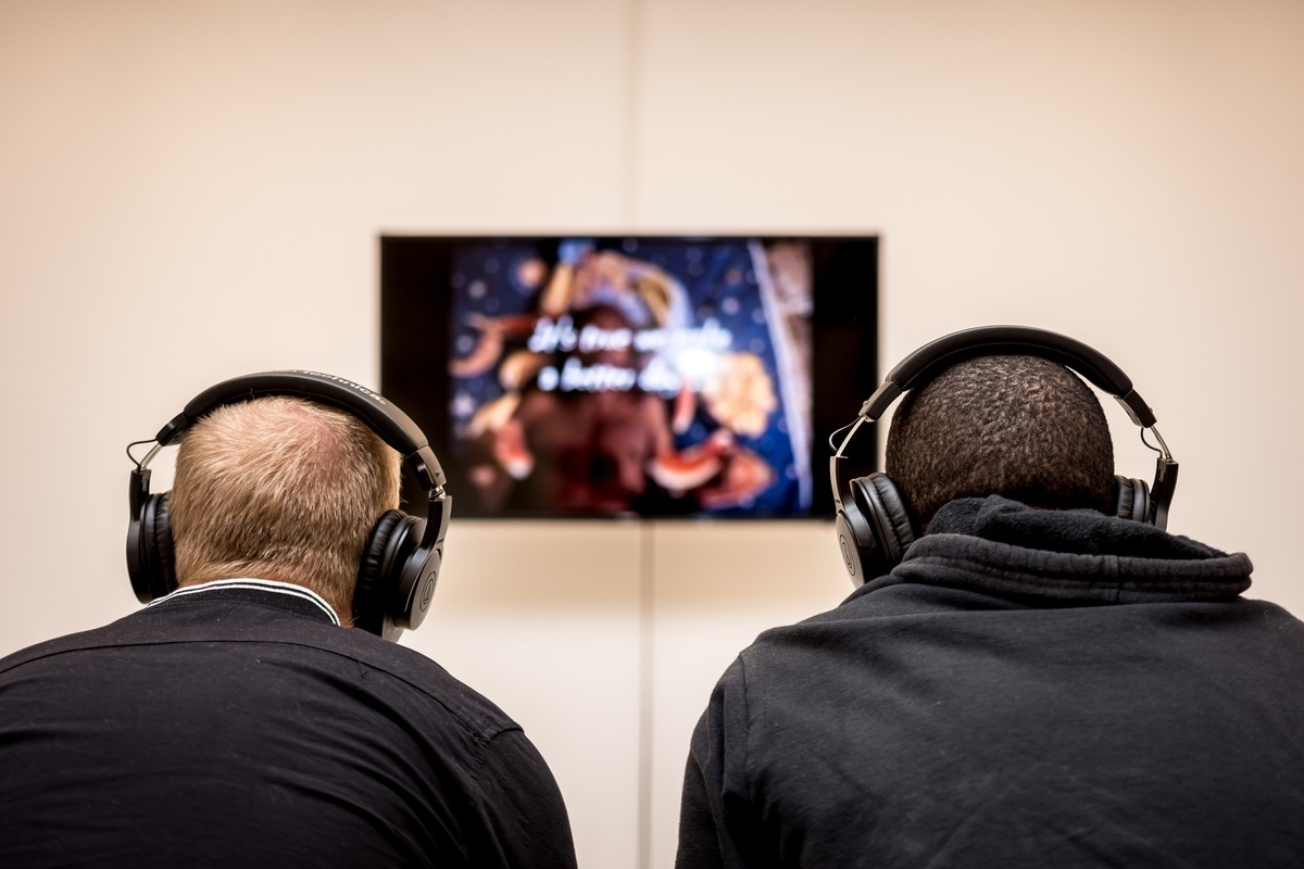 Event photograph from the opening of the You & I exhibition in A4’s Gallery. At the back, Goddy Leye’s video ‘We are the world’ is playing on a wall-mounted screen. At the front, two attendees are viewing the work wearing the accompanying headphones.
