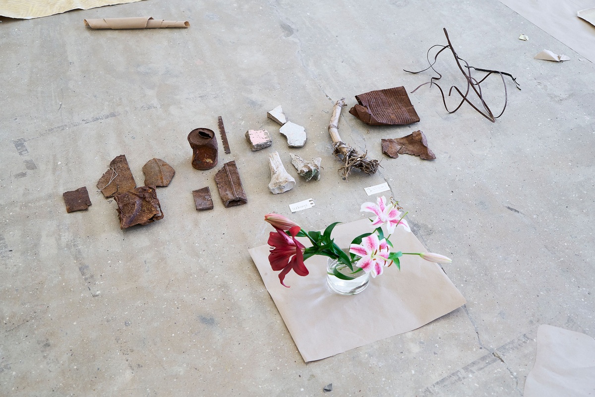 Process photograph of Asemahle Nltonti’s working space during the 2nd rendition of ‘Parallel Play’ in A4’s Gallery. A glass vase with flowers sits next to a collection of found objects that line the concrete floor, including fragments of rusted metal, painted cement and bone.
