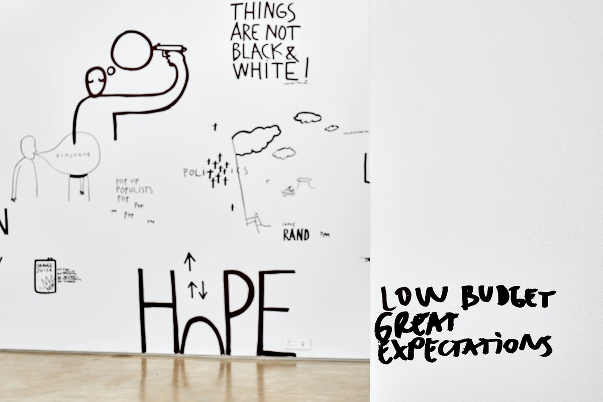 Installation photograph from Dan Perjovschi’s ‘The Black and White Cape Town Report’ exhibition in A4’s Gallery that shows black felt pen marker drawings on white walls. On the right, the corner of the wall features the phrase ‘low budget’ above the phrase ‘great expectations’. On the left, the wall reads ‘things are not black and white!’
