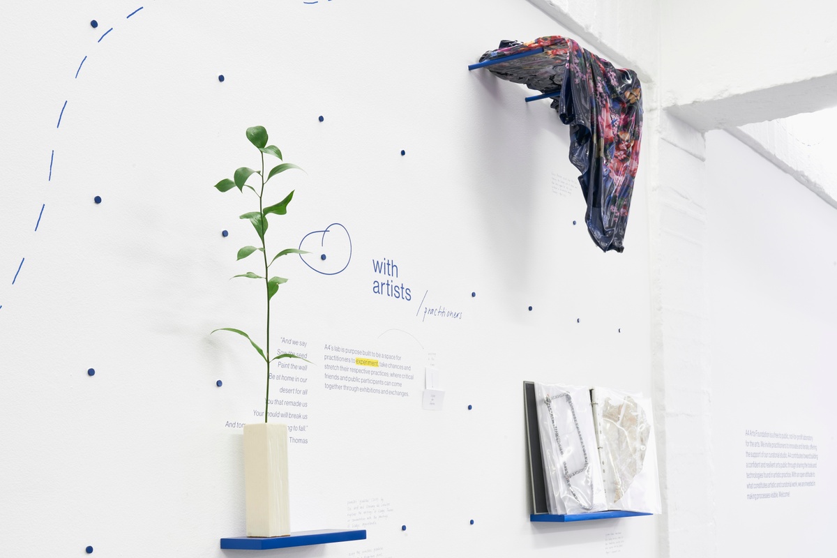 Installation photograph of the A4 About Wall shows the phrase 'with artists/practitioners' described on a white wall in blue. Above, Kevin Beasley's untitled housedress and resin sculpture is mounted on blue dowels. On the right, a folder with process objects by Igshaan Adams. On the left, a branch with leaves in a vase.
