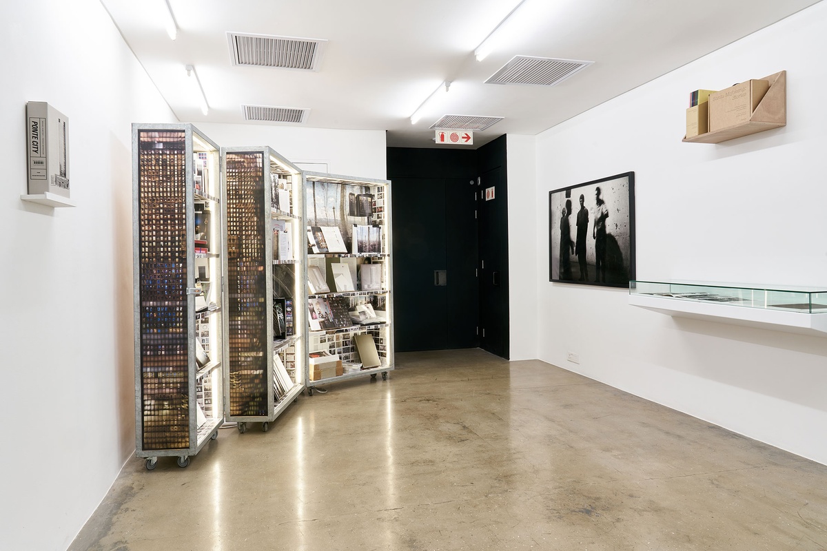 Installation photograph from the Photo Book! Photo-Book! Photobook! exhibition in A4’s Gallery. On the left, Mikhael Subotzky and Patrick Waterhouse’s cabinet installation of the 'Ponte City' archive, and their book ‘Ponte City’ sitting on a wall-mounted shelf. On the right, Santu Mofokeng’s framed monochrome photographic print ‘Concert at Sewefontein, Bloemhof’ is mounted on the wall, alongside a glass-covered display shelf and a wooden display shelf.
