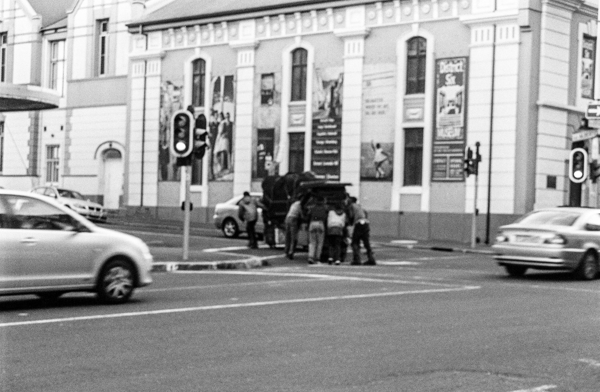 Monochrome event photograph from ‘Contact’, a collaboration between A4’s proto~ museum shop and Oath Magazine, taken by a participant. A group of individuals gather around some sort of conveyance on the corner of Buitenkant and Albertus Street.
