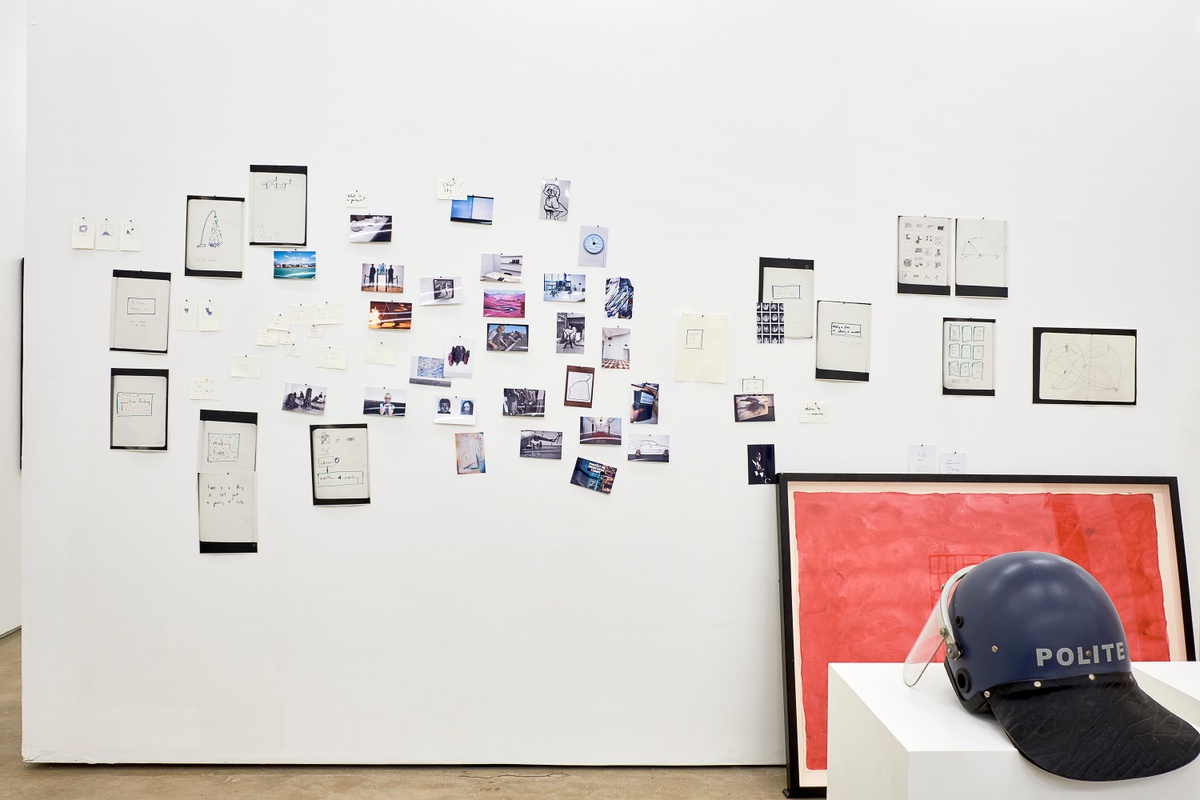 Installation photograph from the 2018 rendition of ‘Parallel Play’ in A4’s Gallery. At the back, photocopies and photographic prints line a moveable gallery wall, with Moshekwa Langa’s gouache and pencil work ‘I Am So Sorry (Red)’ leaning against the wall. On the right, a helmet from Christian Nerf’s ‘Polite Force’ project sits on a white plinth.
