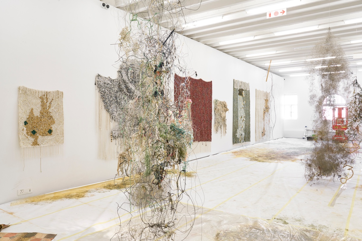 Process photograph from ‘Open Production’, Igshaan Adams’ hybrid studio/exhibition in A4’s Gallery. On the left, a row of Adam’s mixed media tapestries line the gallery wall. On the right, the floor is covered in paper with drawing marks, with Adams’ mixed media wire sculptures are suspended from the ceiling above it.
