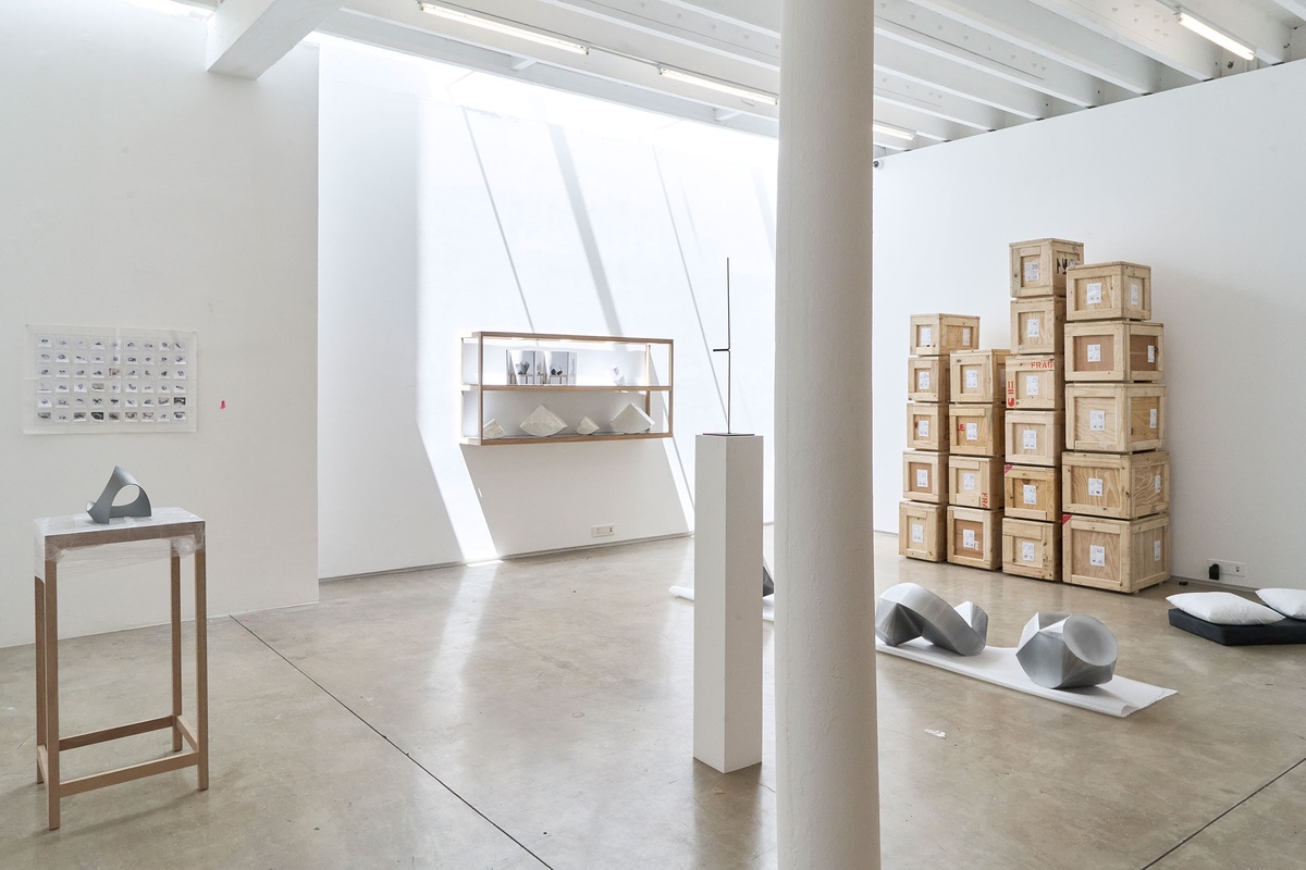 Process photograph of Kyle Morland’s working space during the 2nd rendition of ‘Parallel Play’ in A4’s Gallery. On the right, several wooden crates are stacked against the gallery wall. In the middle, a wall mounted wooden shelf with photographs and sculptural objects. On the left, a wooden stand with a small sculptural object.
