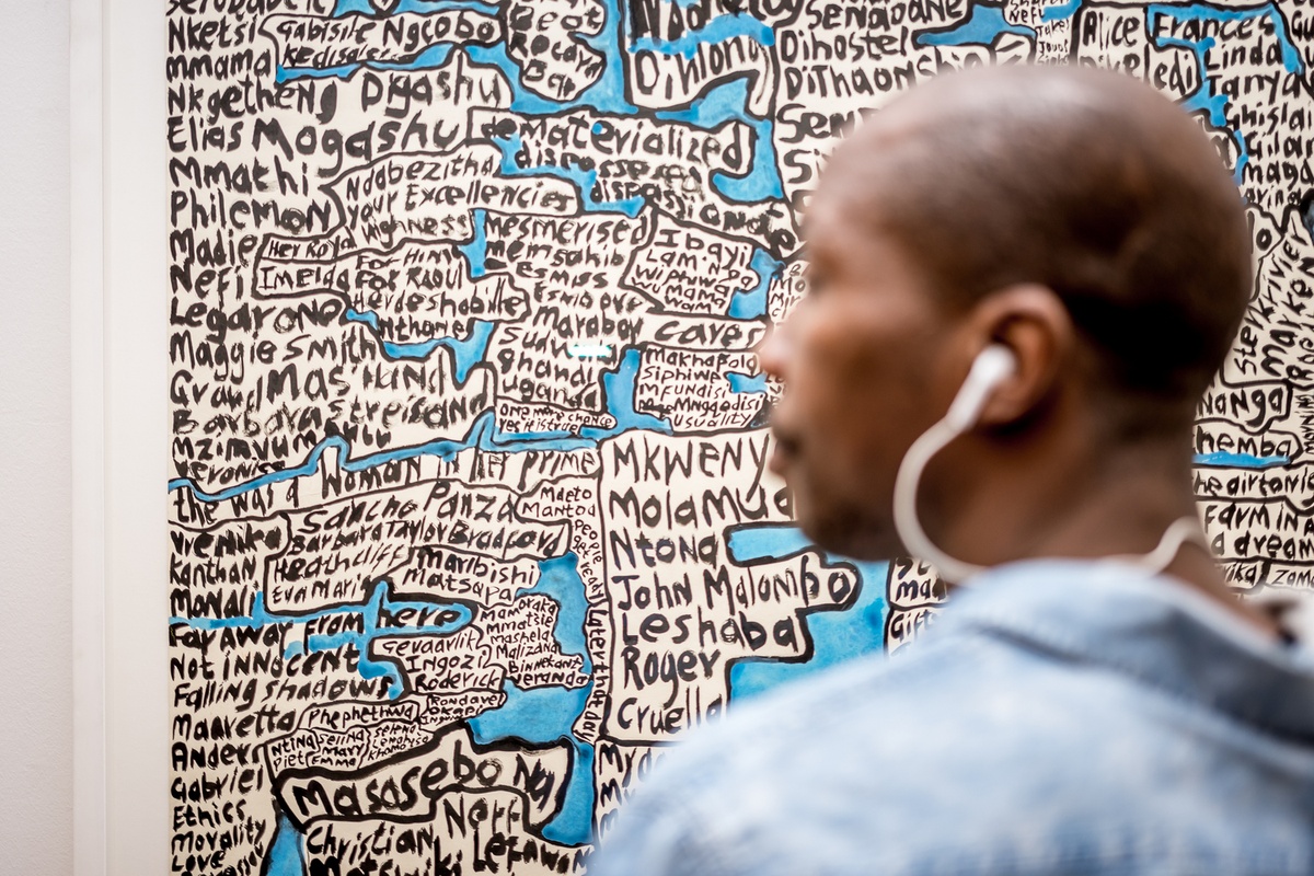 Event photograph from the opening of the You & I exhibition in A4’s Gallery. At the back, Moshekwa Langa’s mixed-media work ‘Mkwenyana (Son in Law)’ is mounted on the gallery wall. At the front, an attendee wearing an electronic earpiece.
