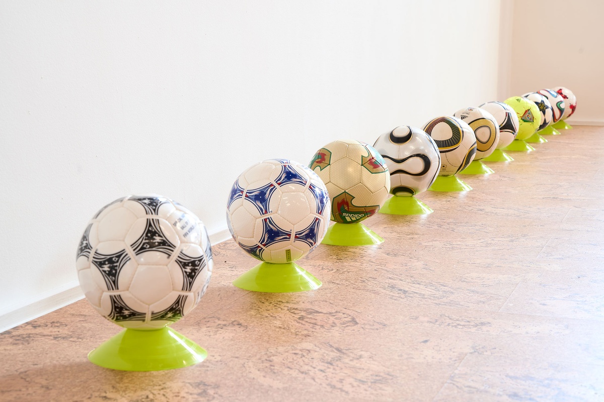Installation photograph from the 2022 rendition of ‘Exhibition Match’ on A4’s second floor. Soccer balls sit on plastic stands in front of a white wall.
