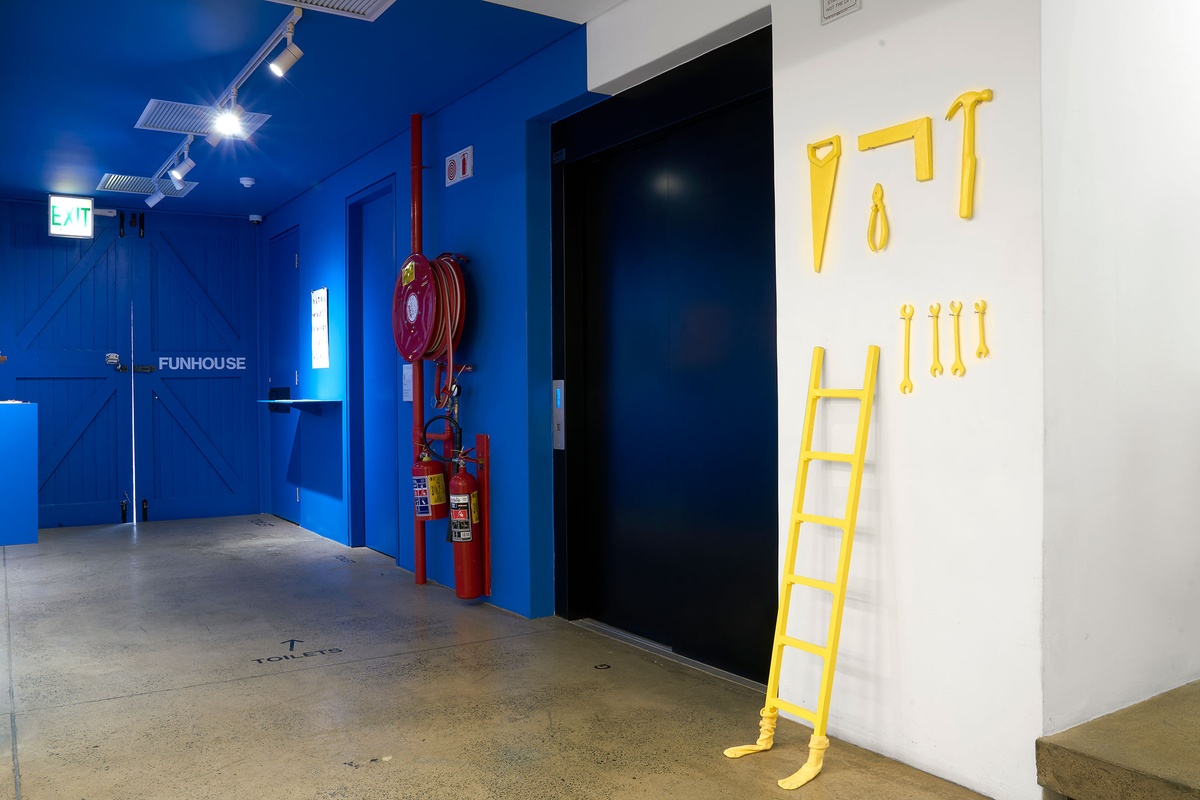 Installation photograph from the ‘Funhouse’ exhibition in A4’s Goods project space that features Luca Evans’ ‘Object Font’ project. On the left, a blue shelf sits under a poster mounted on a blue wall. On the right, yellow sculptures of various tools are mounted on a white wall, along with a sculpture of a ladder that stands on the floor and is fitted with yellow socks.
