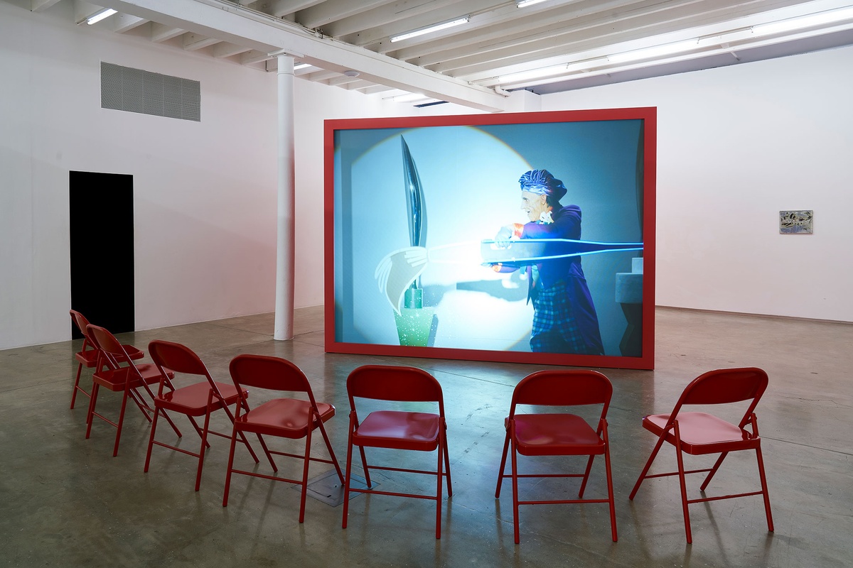 Installation photograph from the 'A Little After This' exhibition in A4 Arts Foundation's gallery that shows Alex Da Corte's video installation 'ROY G BIV'. At the back, a large red wooden box with a back-projected screen plays Da Corte's video. At the front, 7 red powder-coated viewing chairs are arranged in an arch.
