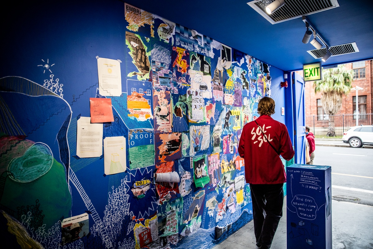 Event photograph from the opening of ‘Disruption’, Hanna Noor Mahomed’s residency in A4’s Goods project space. An attendee walks along a wall mural that consists of drawings and collaged material from Khanya Mashabela’s ‘Social’ archive.
