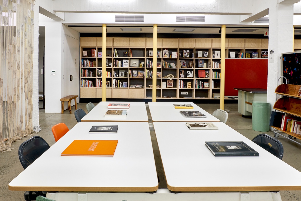 Installation photograph from the ‘Picture Theory’ exhibition in A4’s Gallery that depicts an installation in A4’s Library. At the back, A4’s Library shelves line the wall. At the front, a selection of David Goldblatt’s photo-books are arranged on tables with with seating.
