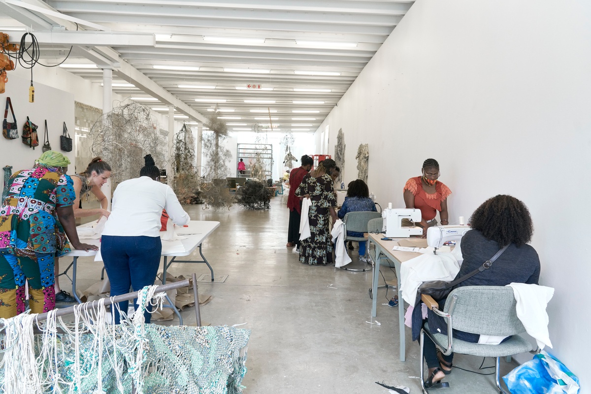 Process photograph from ‘Open Production’, Igshaan Adams’ hybrid studio/exhibition in A4’s Gallery. On the left, two individuals and designer Nicola West handle fabric on a white table. On the right, numerous individuals work at tables set up with sewing machines.
