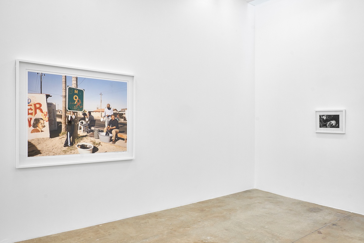 Installation photograph from the ‘Picture Theory’ exhibition in A4’s Gallery. On the left, David Goldblatt’s photograph ‘At Kevin Kwanele’s Takwaito Barber. Lansdowne Road, Khayelitsha, Cape Town in the time of AIDS’ is mounted on the gallery wall. On the right, Goldblatt’s monochrome photograph ‘Sitting next to each other, strangers often become intertwined in sleep’ is mounted on the gallery wall.
