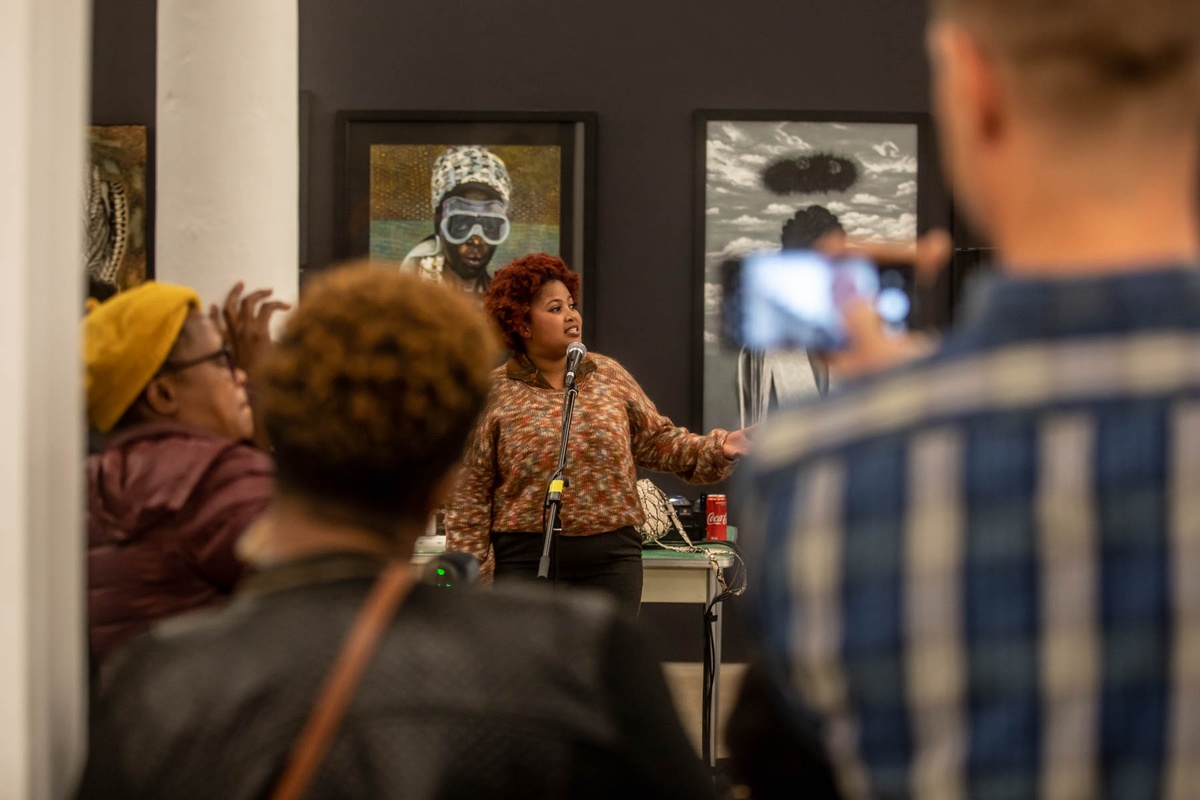 Event photograph from ‘Finissage: Ikhono LaseNatali’ in A4’s Gallery. At the front, attendees for the even are standing. In the middle, vocalist Michelle Fani performs with a microphone stand. At the back, Mthobisi Maphumulo’s oil pastel drawing ‘Somnyama Ngoyama’ and Mduduzi Dzanibe’s acrylic painting ‘Somnyama Ngoyama’ are mounted on the gallery wall.
