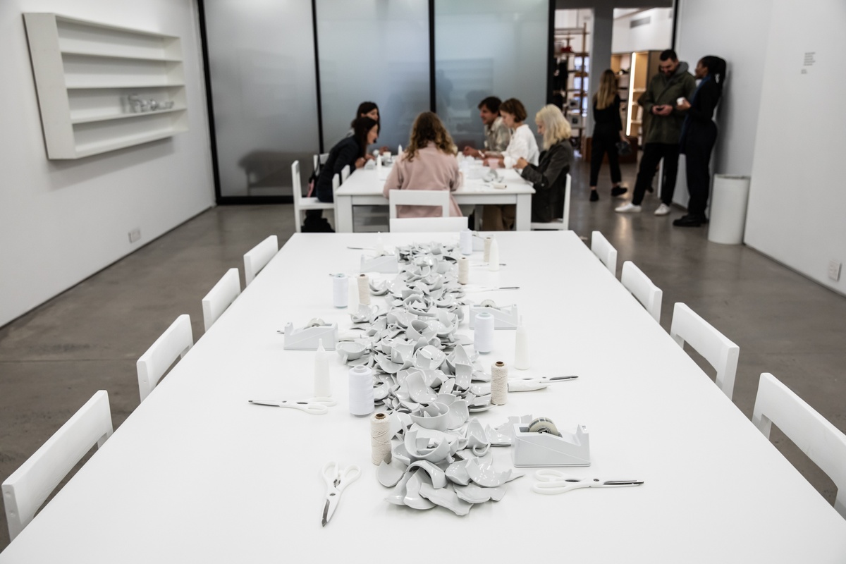 Event photograph from the opening of the Customs exhibition in A4’s Gallery that shows Yoko Ono’s installation ‘MEND PIECE, A4 Arts Foundation, Cape Town version’ in A4’s ground-floor Reading Room. In the middle, participants are seated around a white table interacting with the installation. On the left, a white wall-mounted shelving unit holds some sculptural forms produced by the participants.
