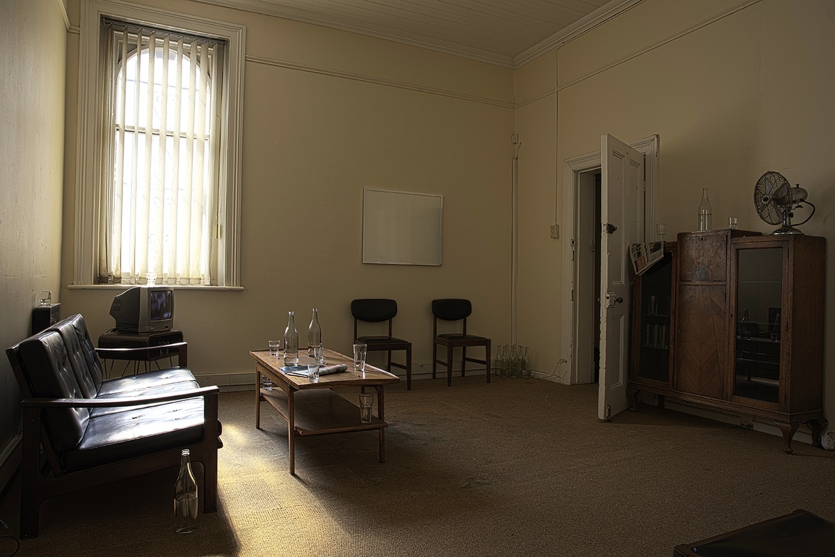 Installation photograph from Gain Maria Tosatti’s offsite exhibition ‘My Hart is so Leeg soon ’n Spieël (My Heart is a Void, the Void is a Mirror)’. On the left, a black couch. In the middle, a wooden coffee table with glass bottles, drinking glasses and printed matter. On the right, a wooden display cabinet. At the back, two chairs at the base of the wall with a blank frame on the wall above it.
