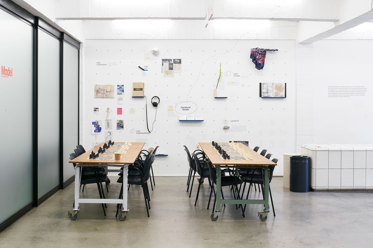 Installation photograph from the launch of Brett Seiler’s chess set in A4’s Proto museum shop that shows the chess tournament setup. Two long wooden tables with chairs host a series of travelling chess sets.
