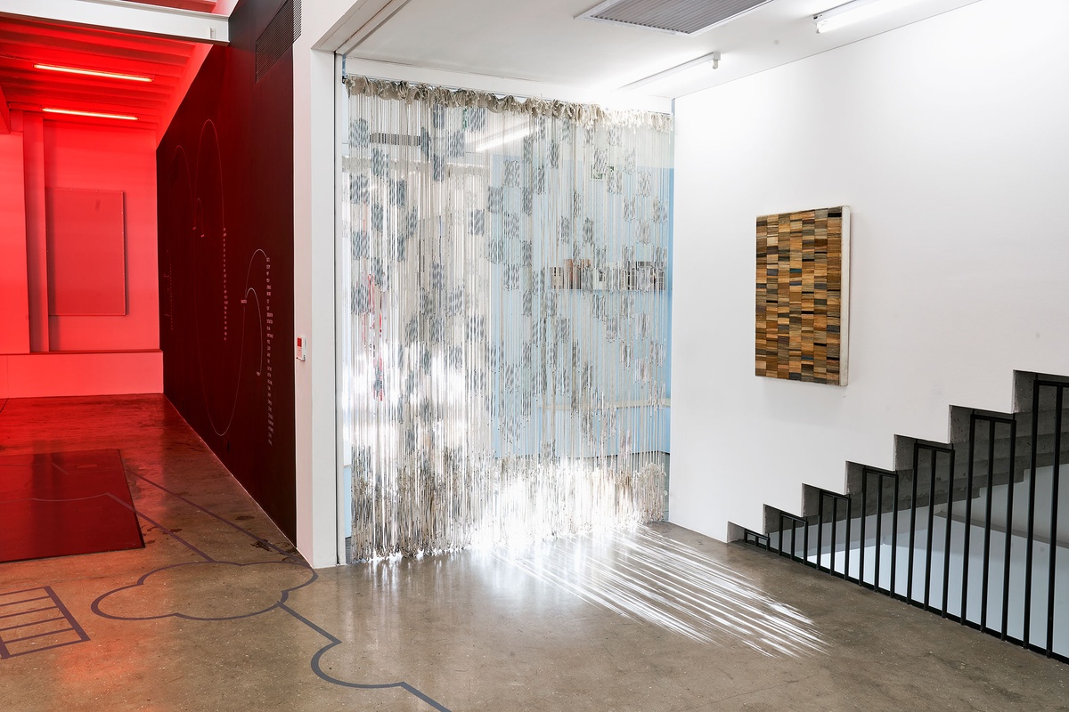 Installation photograph from the Customs exhibition in A4’s Gallery. In the middle, Igshaan Adams’ tapestry ‘Al asma ul Husna’ is suspended from the ceiling. On the right, Willem Boshoff’s cardboard work ‘Blind Cards’ is mounted on a white gallery wall.
