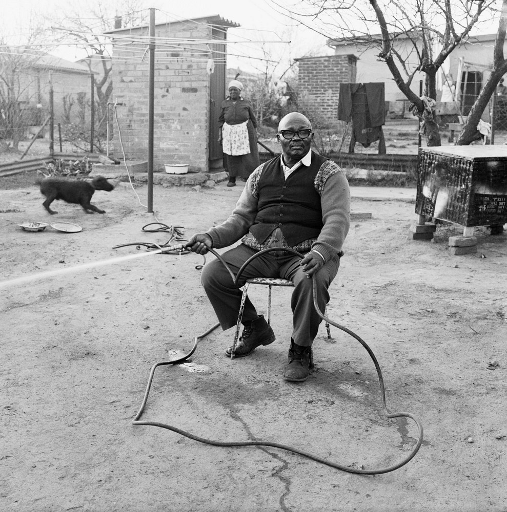 David Goldblatt's black-and-white photograph 'Ephraim Zulu watering his garden, 179 Central Western Jabavu, Soweto. September' shows a man seated on a chair in a yard, holding a hosepipe. In the background is a dog and a woman.
