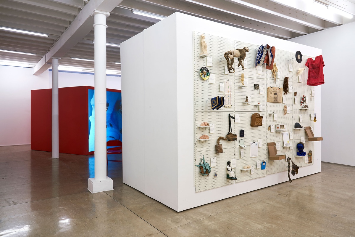 Installation photograph from the 'A Little After This' exhibition in A4 Arts Foundation's gallery. On the right, collected objects from Penny Siopis' 'Will' work are mounted on a white moveable gallery wall through the use of perforated metal panels. On the left, a large red wooden box with a back-projected screen from Alex Da Corte's video installation 'ROY G BIV'.
