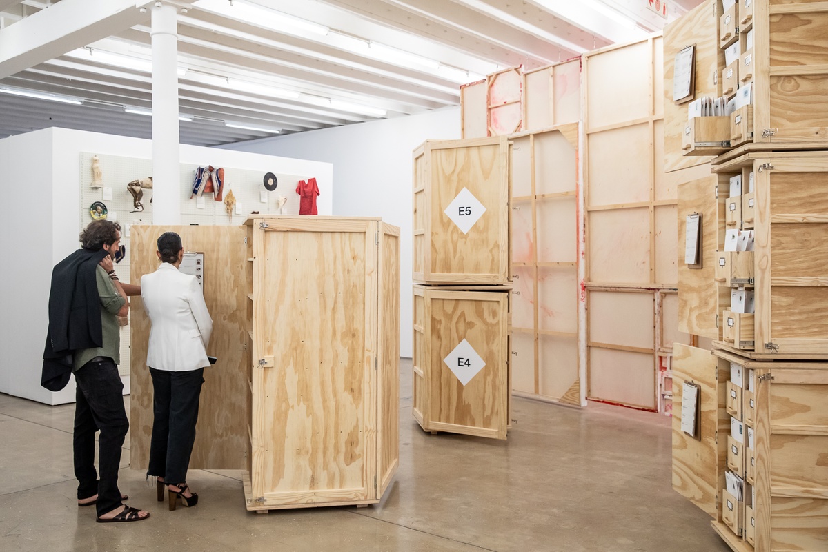 Event photograph from the preview of the 'A Little After This' exhibition in A4 Arts Foundation. At the front, collected objects from Penny Siopis' 'Will' work are stored in envelopes and wooden crates. At the back to the left, collected objects from Penny Siopis' 'Will' work are mounted on a white moveable gallery wall through the use of perforated metal panels. In the back to the right, an installation of Penny Siopis' glue, ink and oil 'Maitland' paintings suspended from the ceiling.
